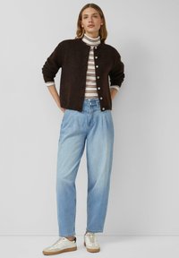 Brązowy dziergany kardigan z okrągłym dekoltem i guzikami, noszony na paskowany golf, zestawiony z jasnoniebieskimi, wysokim stanem dżinsami oraz białymi trampkami.