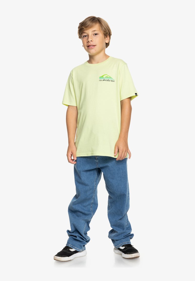 Camiseta de algodón verde claro con un logotipo gráfico en el pecho, combinada con unos vaqueros de mezclilla azules y zapatillas negras con suelas blancas.