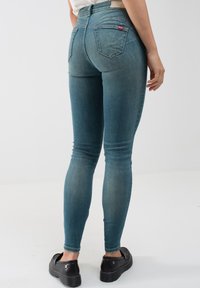 Mėlynos džinsinės liesos kelnės su aukšta juosmens linija, dviem galiniais kišenėmis ir mažyčiu prekių ženklu. Dėvimos su juodais platforminiais batais.