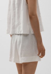 Top alb din in foiță fără mâneci, cu fante pe laterale, purtat cu pantaloni scurți asortați. Textură moale și croială lejeră. Detalii discrete ale cusăturii.