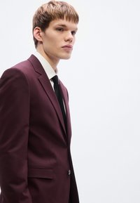 Μπορντό κοστούμι με εφαρμοστή γραμμή, με μονό κουμπί κλεισίματος, πέτα με νύχι και μαύρη γραβάτα πάνω από λευκό πουκάμισο.