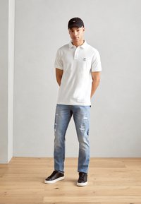 Hvid polo-shirt med korte ærmer og et lille logo, parret med lyseblå slidte jeans og sorte sneakers med hvide såler.