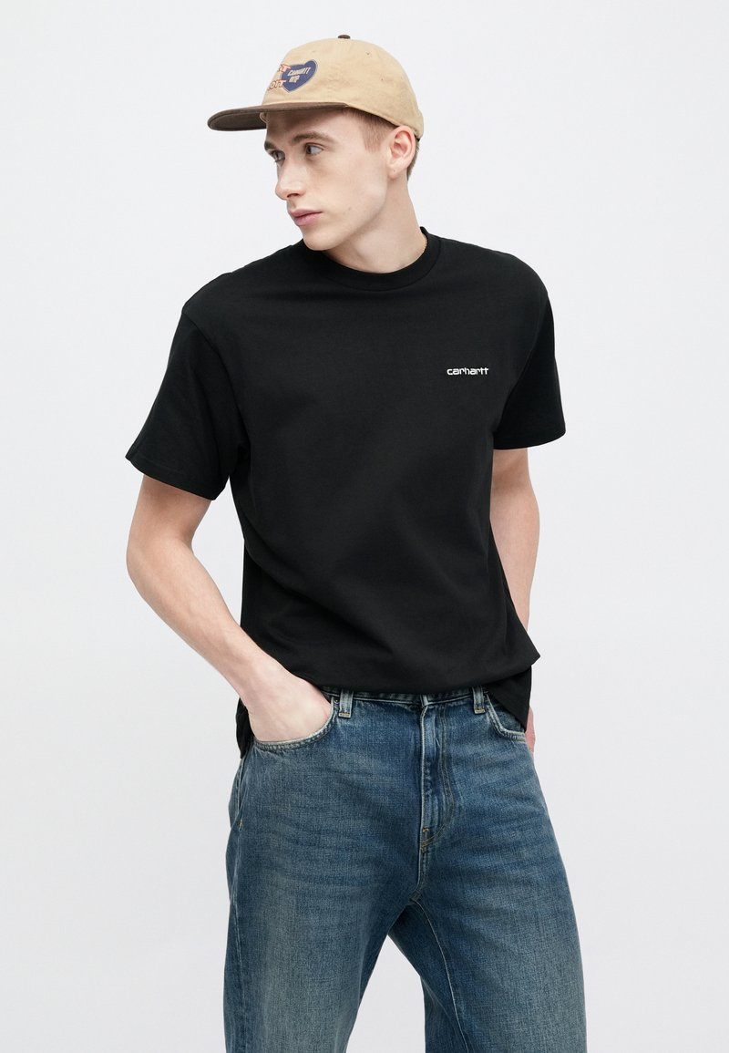 Black cotton t-shirt with short sleeves, crew neck, and front logo. Paired with faded blue denim jeans and a beige cap with a logo.