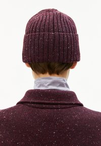 Een bordeauxrood geribbeld gebreid mutsje met gespikkelde patronen, gedragen over een grijze kraag. De textuur lijkt zacht en warm. Het zicht toont de achterkant.