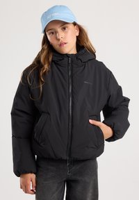 Black puffy jacket with a high collar, front zipper, and side pockets. Paired with a light blue cap featuring embroidered text.