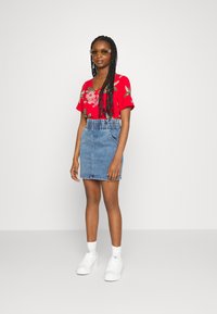 Top floral vermelho com mangas curtas, saia lápis de denim com cintura franzida e ténis brancos. Modelo está de pé contra um fundo liso.