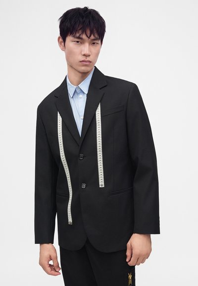 Homme portant une veste de costume noire et une chemise bleu clair avec un mètre ruban drapé autour du cou sur un fond uni.