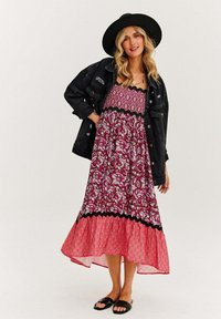 Maxi sukienka w purpurowy kwiatowy wzór, z czarnym falbaniastym wykończeniem i różowym dolnym brzegiem, założona pod czarną dżinsową kurtką i kapeluszem. Czarne sandały.