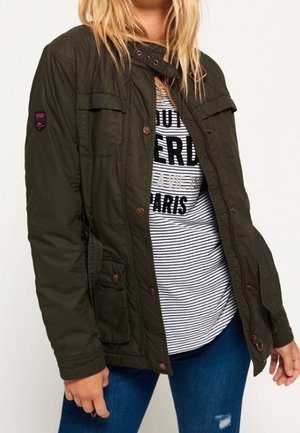 Olivengrøn, quiltet jakke med knaplukninger og brystlommer. Har en høj krave og en glat tekstur, båret over en stribet skjorte.