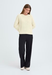 Maglione a maglia beige chiaro con scollo rotondo, maniche lunghe e texture a coste, abbinato a pantaloni neri larghi e sneakers bianche.