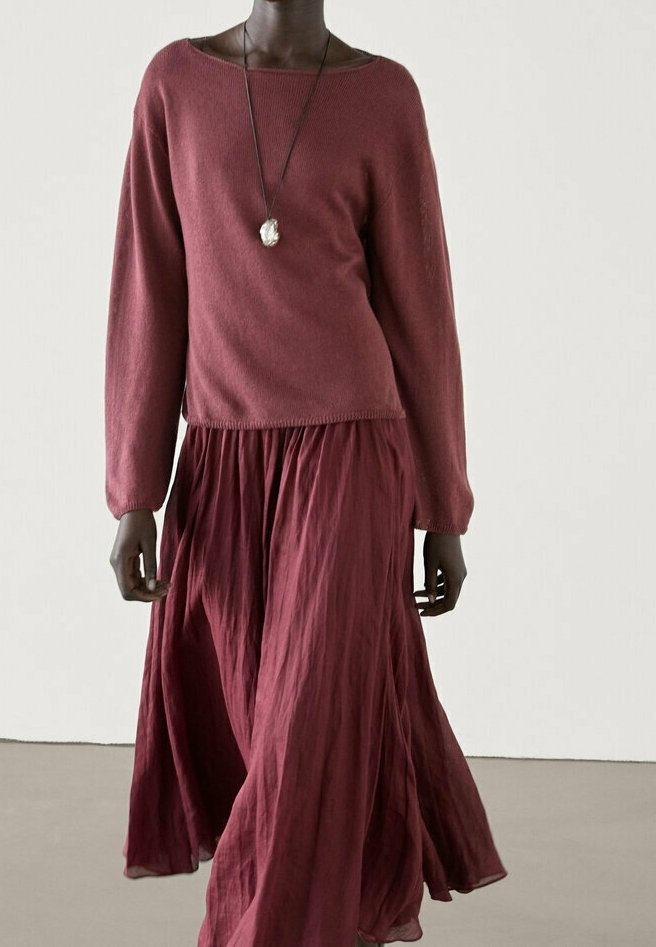 Pitkä viininpunainen neulepaita leveällä pääntiellä, yhdistettynä virtaavaan viininpunaiseen maksihameeseen. Kangas näyttää pehmeältä ja teksturoidulta, korostaen kerroksia.