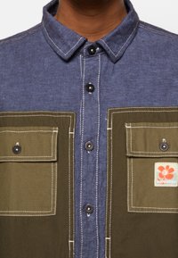 Chemise bleu et olive boutonnée avec deux grandes poches avant, coutures blanches, et un petit logo fleur orange sur la poche droite.