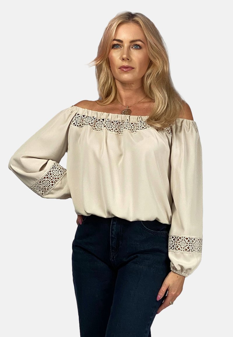 Beżowa bluzka typu off-the-shoulder z koronkowymi detalami wzdłuż dekoltu i mankietów, charakteryzująca się luźnymi rękawami i gładką fakturą.