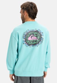 Turquoise sweatshirt with long sleeves, featuring a large, colorful graphic on the back of a globe and logo. Soft cotton texture.