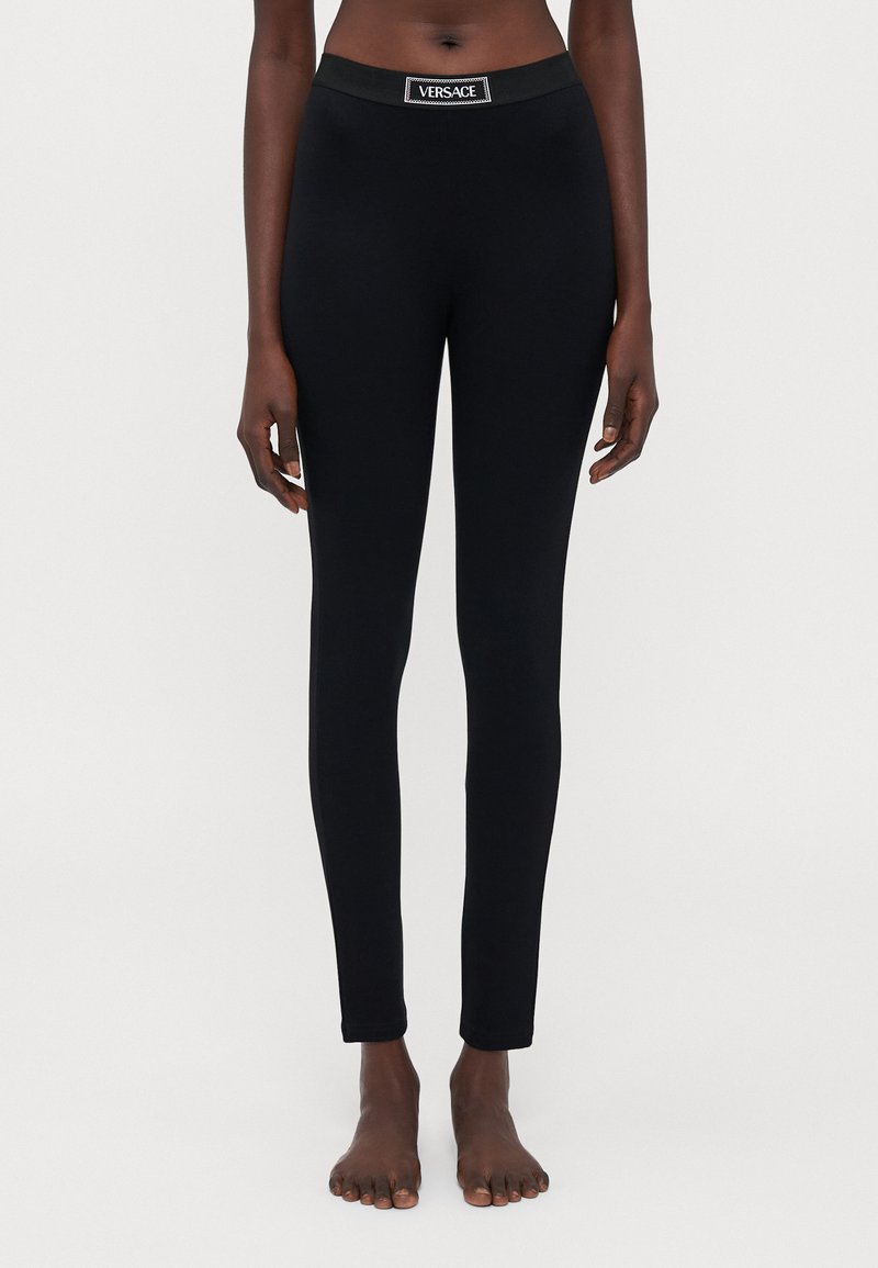 Colanți negri cu un design ajustat și textură netedă. Acestea au un waistband elastic cu brand cu logo-ul "VERSACE" în față.