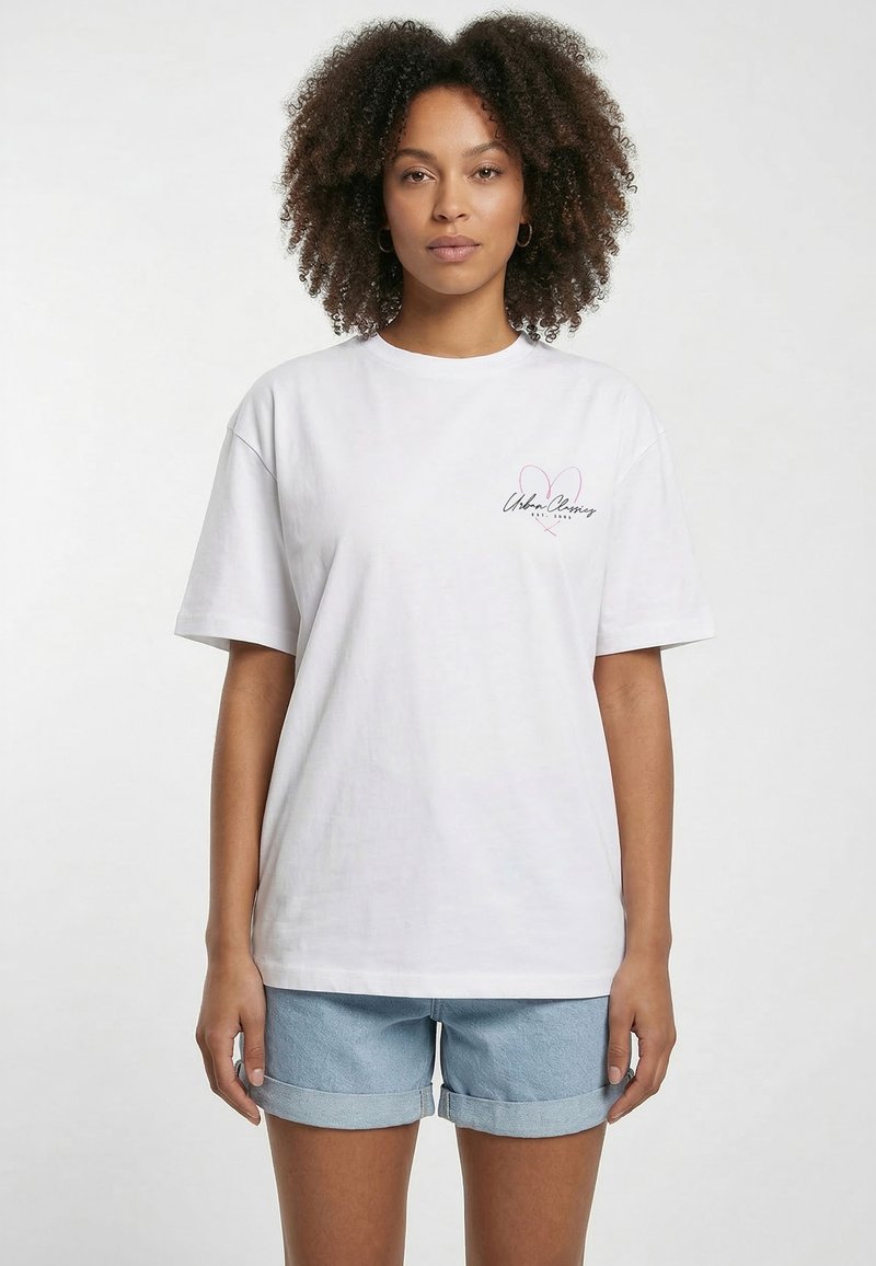 Femme aux cheveux bouclés portant un t-shirt blanc avec un petit cœur et un texte sur la poitrine, ainsi qu'un short en denim bleu clair roulotté.