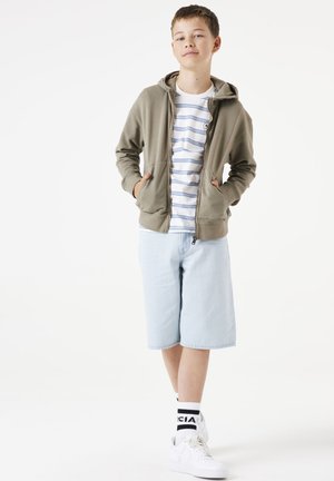 Veste à capuche zippée olive sur un t-shirt rayé blanc et bleu, assortie à un short en jean bleu clair et des baskets blanches avec des chaussettes noires.