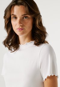 Λευκό ριμπ μπλουζάκι με ψηλό λαιμόκοψη και κοντά μανίκια με βολάν. Διαθέτει διακριτικό λογότυπο στο στήθος. Μαλακή υφή.