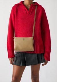 En person i en röd stickad tröja och rutiga veckkjolar, som bär en beige läder crossbody-väska med en metallsymbol.