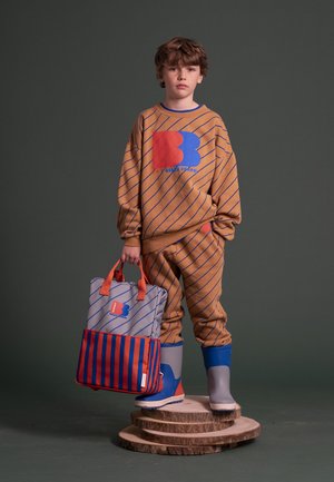 Garçon en tenue de survêtement marron rayée et bottes gris-bleu tenant un sac rayé assorti, debout sur des rondins de bois empilés sur fond gris.