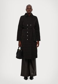 Melna izšūta mētelis ar apkakli, pogu priekšpusi un jostu. Nēsāta virs tumšām palazzo biksēm un turēta melna rokassoma. Gluda tekstūra, minimāls dizains.