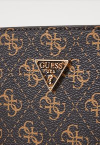 Juodas tekstūruotas medžiagos rankinukas, turintis pakartotinį rusvą raštą ir auksinę trikampę logotipo plokštelę su tekstu "GUESS USA".