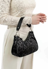 Must ja hall leopardimustriga käekott läikivate naelakeste, nahase detailide ja eemaldatava õlarihmaga, mida hoiab käes inimene sädelevas topsis.