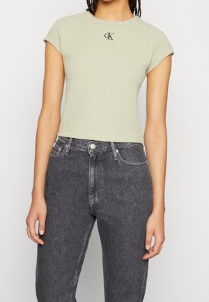 Osoba ubrana w jasnobeżowy krótki t-shirt z logo cK oraz czarne dżinsy z wysokim stanem, stojąca na jednolitym tle.