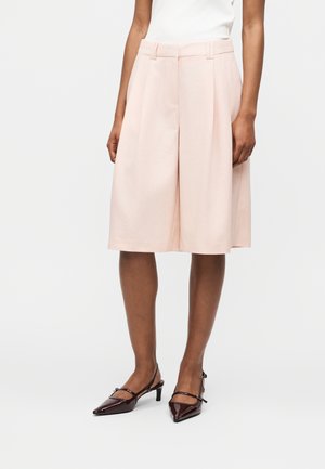 Persoon draagt lichtroze wijde knielange shorts, wit topje en donker bordeauxrode lage hak puntige schoenen met enkelbandjes.