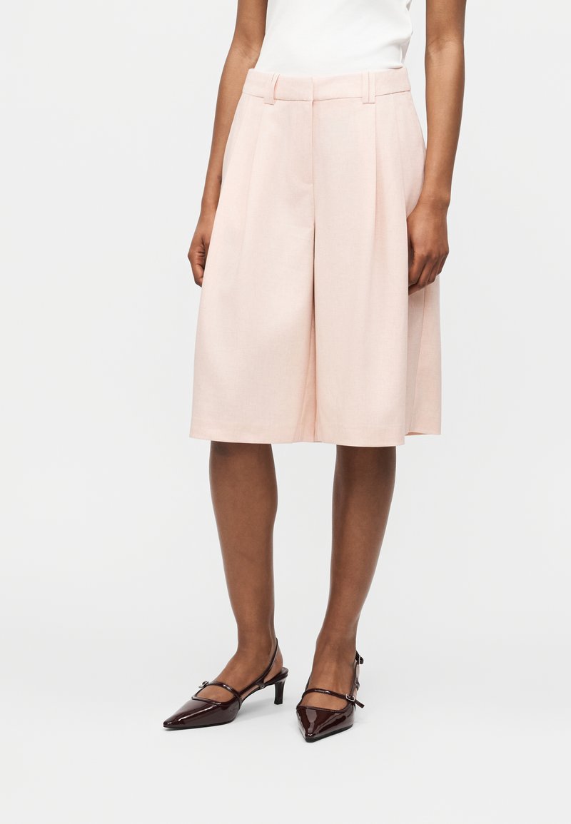 Άτομο φορέσει ανοιχτό ροζ, φαρδιά σορτς μέχρι το γόνατο, λευκό τοπ, και σκούρα βουργουνδί χαμηλά τακούνια μυτερά παπούτσια με λουράκια στο αστράγαλο.