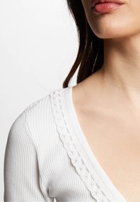 Biały prążkowany top z głębokim dekoltem w kształcie litery V ozdobionym koronką wokół kołnierza, o gładkiej fakturze.