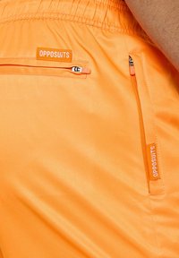 Oranžinės šortai pagaminti iš lygaus, blizgaus audinio su elastinga juosmens juosta, turi užtrauktuku uždaromas kišenes ir "OPPOSUITS" etiketes.