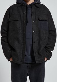 Giacca in lana nera con finitura testurizzata, dotata di due tasche sul petto, bottoni a pressione e una tasca a toppa nera. Indossata sopra un hoodie blu navy.