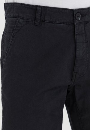 Schwarze Baumwollhose mit glatter Textur, ausgestattet mit einem Knopfverschluss, Gürtelschlaufen und Seitentaschen. Nahaufnahme des Taillenbands und der Nähte.