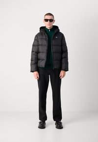 Zwarte pufferjack met een capuchon, gecombineerd met een donkergroene ritsjack, zwarte broek en zwarte schoenen, gedragen door een model.