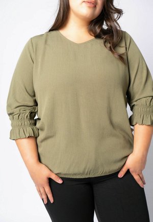Mujer con blusa verde oliva con mangas 3/4 fruncidas y pantalones negros, de pie con las manos apoyadas en las caderas sobre un fondo blanco.
