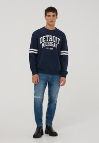 Granatowa bluza z napisem "DETROIT MICHIGAN EST. 1982", białe paski na rękawach, zestawiona z przetartymi niebieskimi dżinsami i czarnymi butami.