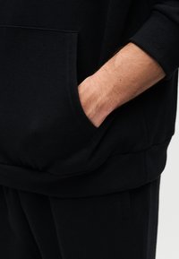 Μαύρο φούτερ με τσέπη καγκουρό, ριμπ μανσέτες και τελείωμα. Το υλικό φαίνεται απαλό με λεία υφή, και η σχεδίαση είναι με χαλαρή εφαρμογή.