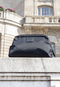 Musta suorakaiteen muotoinen vetoketjullinen pussukka, jossa pieni neliön muotoinen logo, asetettuna kivihyllylle historiallisesta beigevärisestä kivirakennuksesta edessä.