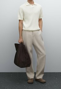 Homme portant un pull à manches courtes crème à côtes et un pantalon beige, tenant un grand sac en cuir souple marron foncé d'une main.