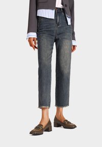 Jeans in denim scuro con taglio dritto e orlo sfrangiato, indossati con una camicia celeste con colletto e scarpe con tacco quadrato marroni.