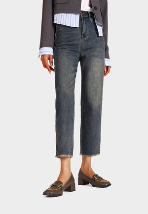 Jeans in denim scuro con taglio dritto e orlo sfrangiato, indossati con una camicia celeste con colletto e scarpe con tacco quadrato marroni.