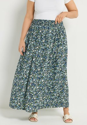 Jupe à imprimé floral bleu, blanc et vert portée avec un haut blanc et des sandales blanches à enfiler, posant devant un fond uni.