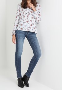 Chemise blanche à manches longues avec motif floral, associée à un jean bleu ajusté et des bottines noires. Textures subtiles et coupe ajustée évidentes.