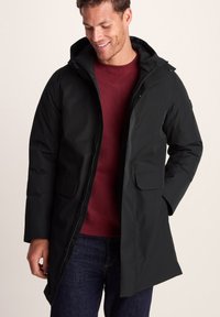 Man wearing black hooded winter coat over a burgundy sweater and dark jeans, smiling and looking down, with one hand in pocket.