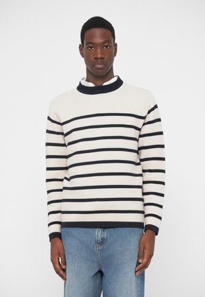 Krém és tengerészkék csíkozott pulóver bordás textúrával, magas gallérral és hosszú ujjakkal. Világoskék farmernadrággal párosítva.