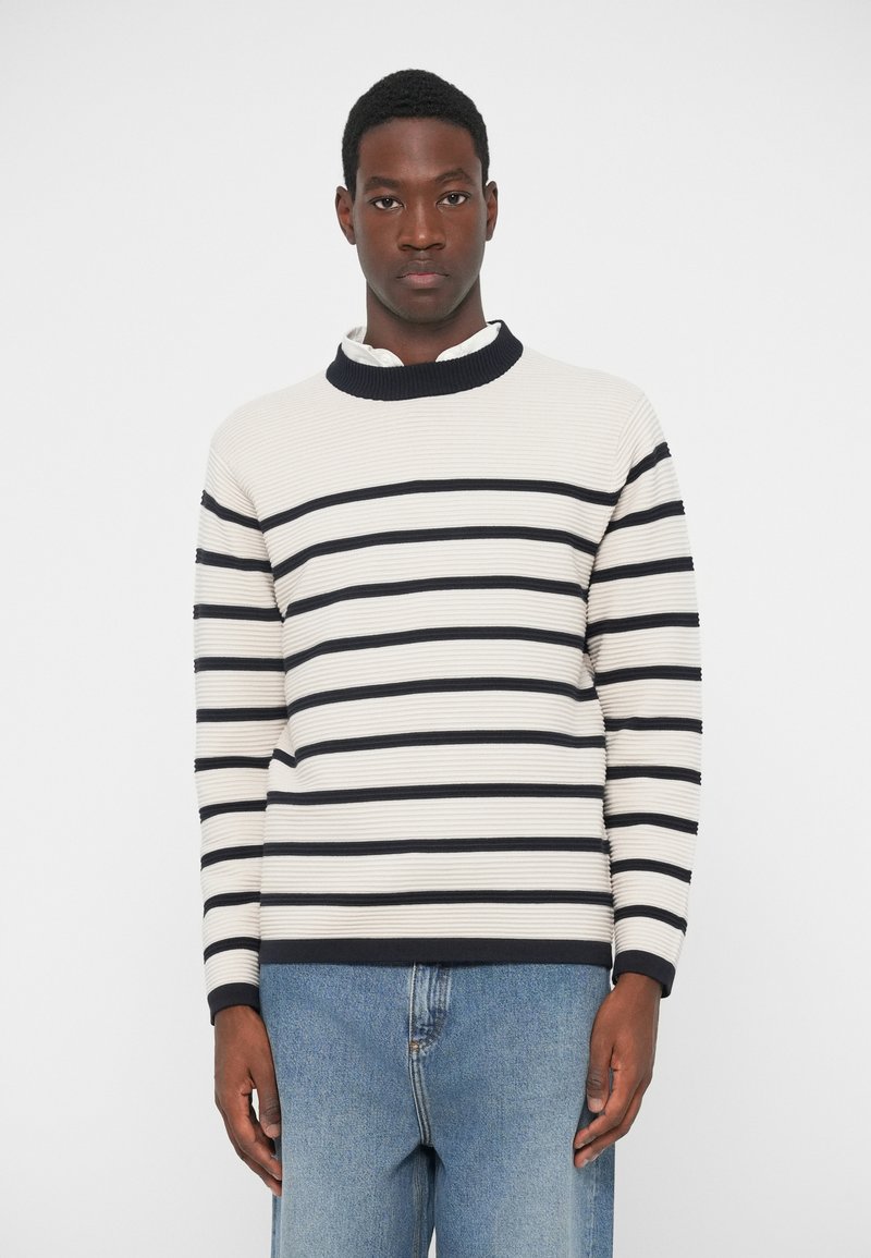 Krema i tamnoplavi prugasti džemper s rebrastom teksturom, visokim ovratnikom i dugim rukavima. Uparen s svijetloplavim trapericama.