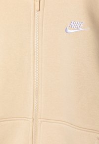Sudadera con cremallera color beige, confeccionada con tejido suave, que presenta un destacado logo bordado de Nike en azul en el pecho y costuras en contraste.