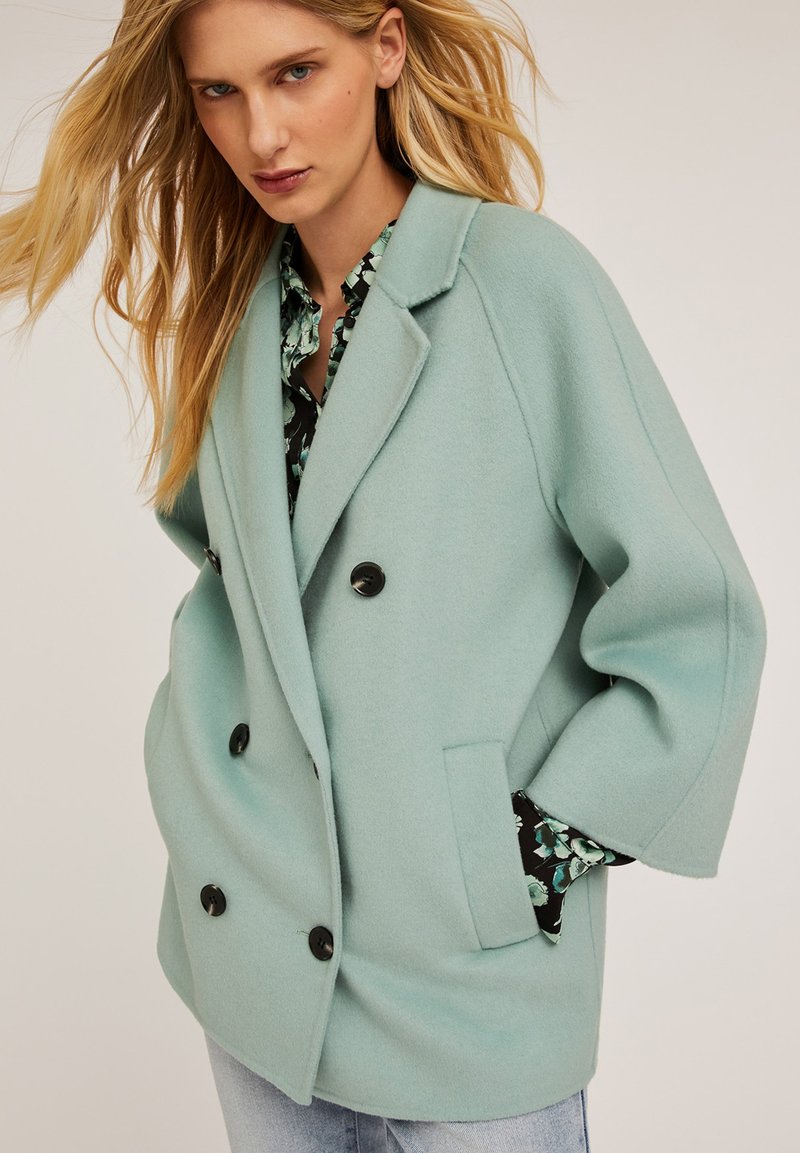 Cappotto in lana verde menta doppiopetto con bottoni neri, maniche ampie e tasche laterali. Foderato con un tessuto a motivi floreali.
