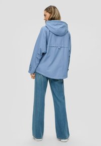 Veste à capuche bleu clair avec une coupe ample, dotée d'une fermeture à boutons et d'un design de rabat au dos. Associée à un jean en denim à jambes larges.